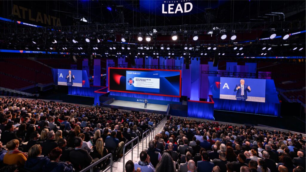 A large conference with a speaker on stage presenting to a packed auditorium. The audience is seated and focused on a large screen displaying graphs and text. Bright lights and a banner reading LEAD are visible above the stage.