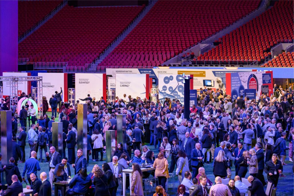 A large, bustling conference with a diverse crowd of people mingling. Booths with signage are set up along the perimeter. Attendees are engaged in conversation, networking, and exploring exhibits in a spacious, brightly lit indoor venue.