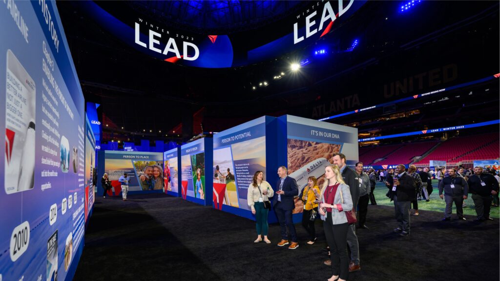 People are observing large, brightly lit informational displays in an indoor stadium setting. The focus is on the exhibition panels with the word LEAD prominently displayed above. The venue is spacious, with a visible section of stadium seating.