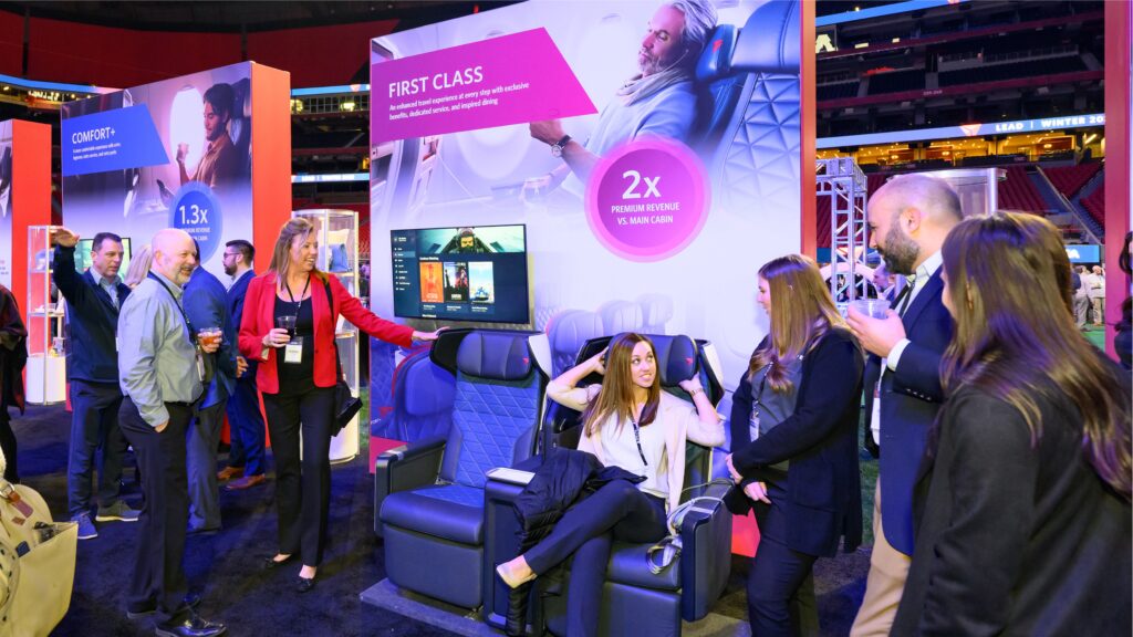 People are gathered around a display booth showcasing first-class airline seats at an exhibition. A woman is sitting in one of the seats, demonstrating its features, while others stand around conversing and observing.