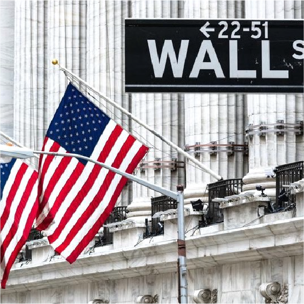 Two American flags waving in front of a building with a Wall Street sign. The building features tall columns and ornate architecture, typical of financial district structures. The scene conveys an essence of the U.S. financial sector.