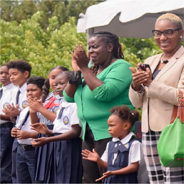 A group of children and two adults are standing under a white tent. The adults, wearing a green top and a plaid blazer, are clapping. The children are in school uniforms, and greenery is visible in the background.