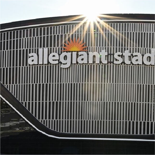 The image shows the exterior of Allegiant Stadium with vertical white lines and the sun casting rays over the structure. The stadiums name is prominently displayed with an orange sunburst logo above the word allegiant.