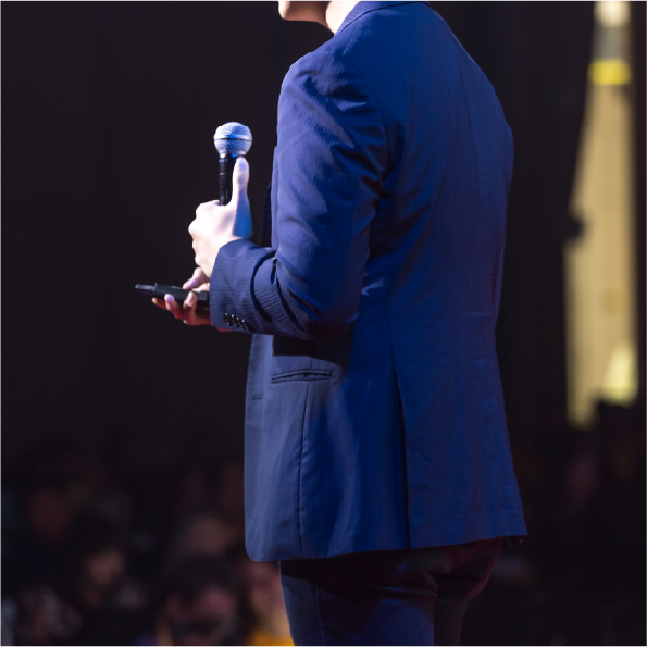 A person in a blue suit holds a microphone and a phone while speaking on stage. The background is dark with blurred audience members. The scene is lit with a spotlight.