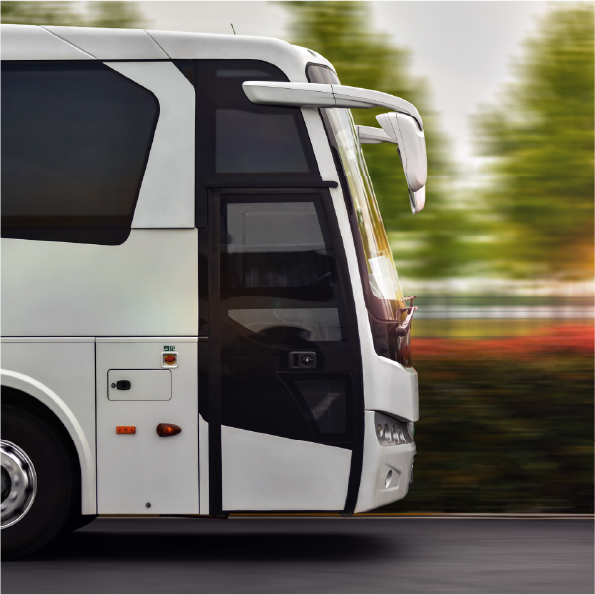 Side view of a moving modern bus on a road, with blurred green trees and red foliage in the background, suggesting motion. The bus has large windows and a sleek, contemporary design.