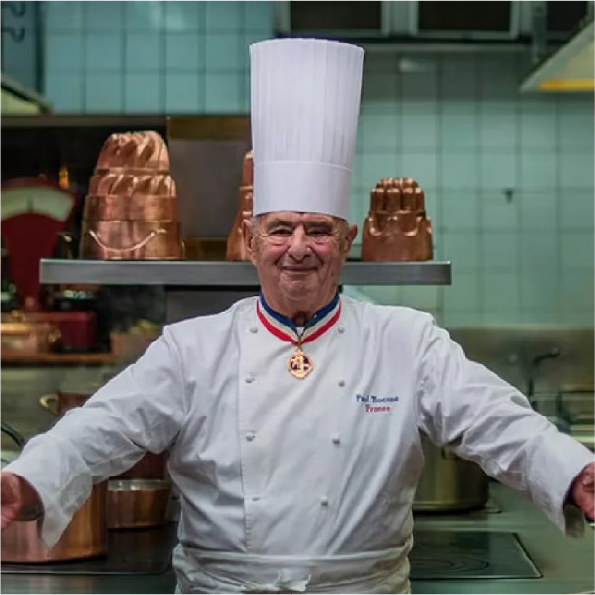 An elderly chef wearing a tall white hat and a white uniform stands in a professional kitchen. He smiles and opens his arms slightly. Copper pots are visible in the background.