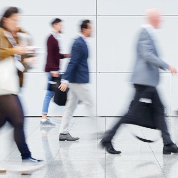Blurred image of several people walking briskly in an indoor setting with a white wall background. They appear to be businessmen and businesswomen, dressed in suits and carrying briefcases or bags. The motion suggests a busy environment.