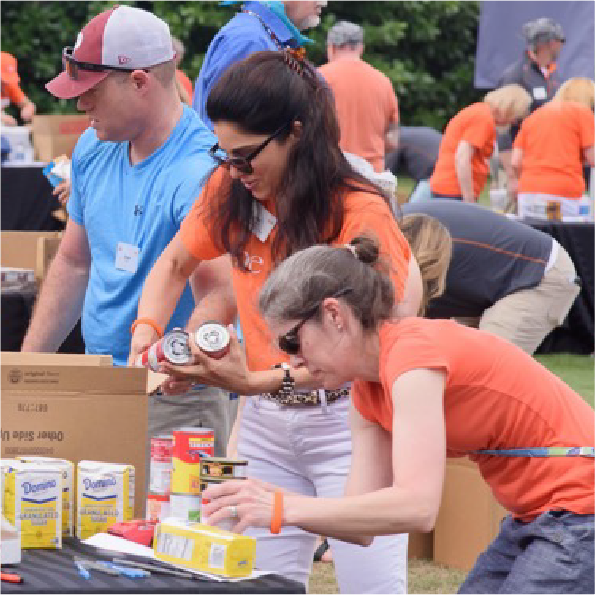 People wearing casual clothes are sorting canned goods and boxed items into cardboard boxes at an outdoor event. Some are wearing orange shirts, and they are focused on organizing the items. There is greenery in the background.