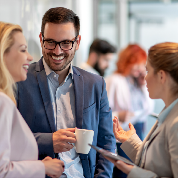 Three professionals are engaged in conversation. A man in a blue suit holds a mug, smiling, while two women, one blonde and one brunette, face him. Blurred figures in the background suggest an office setting.