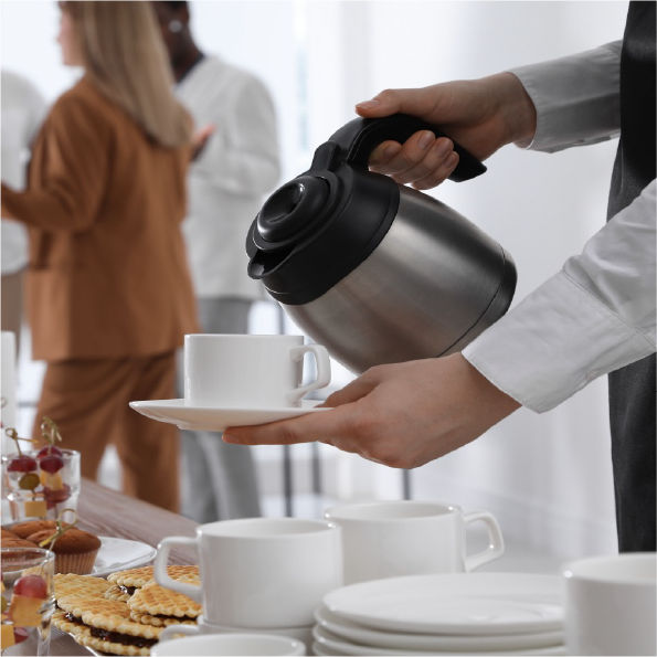 A person holds a metal coffee pot, pouring coffee into a cup on a saucer. In the background, people in business attire are conversing. A table is set with cups, pastries, and decorative desserts.