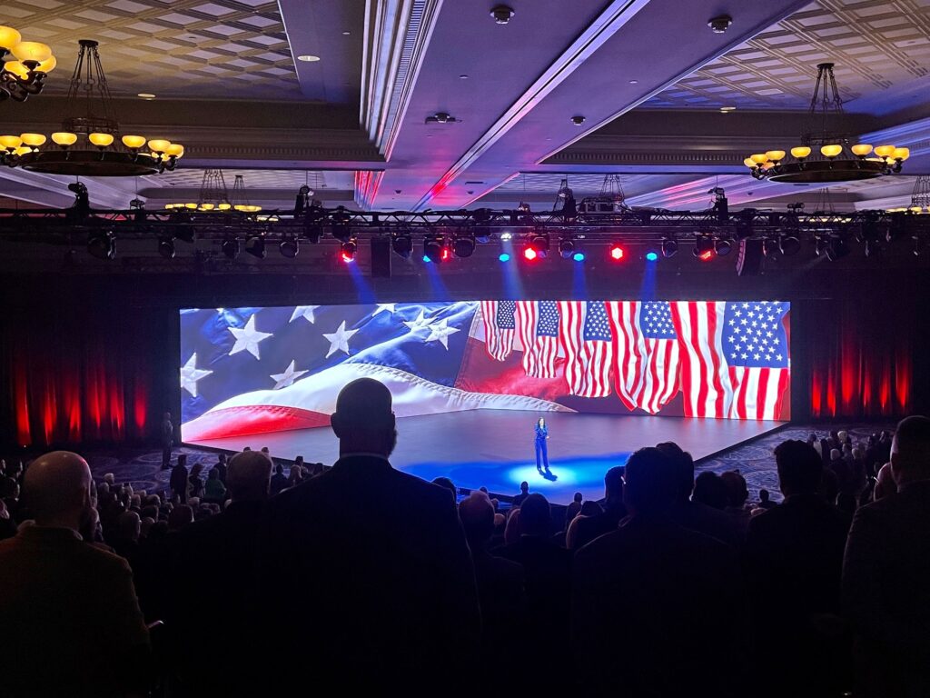 A speaker stands on a stage in front of a large screen displaying multiple American flags. The audience is seated in a dimly lit conference hall with decorative ceiling lights. Red, white, and blue lights illuminate the stage.