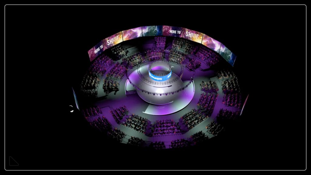 An aerial view of a circular auditorium with tiered seating, illuminated in purple and blue lights. Digital screens display text, and a central circular stage is marked with IMPACT. The venue is filled with an audience.