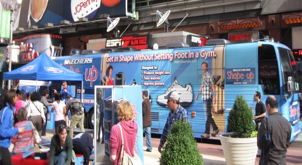 People are gathered around a blue promotional bus with Get in Shape Without Setting Foot in a Gym written on it. The scene is busy, with booths, signs, and a tent nearby. The bus advertises Skechers Shape-ups near a bustling urban area.