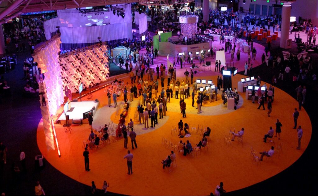 A large convention hall is filled with people. Theres a central circular area with yellow carpeting and various booths. Attendees gather around displays and seating areas. Overhead lights create a colorful ambiance.