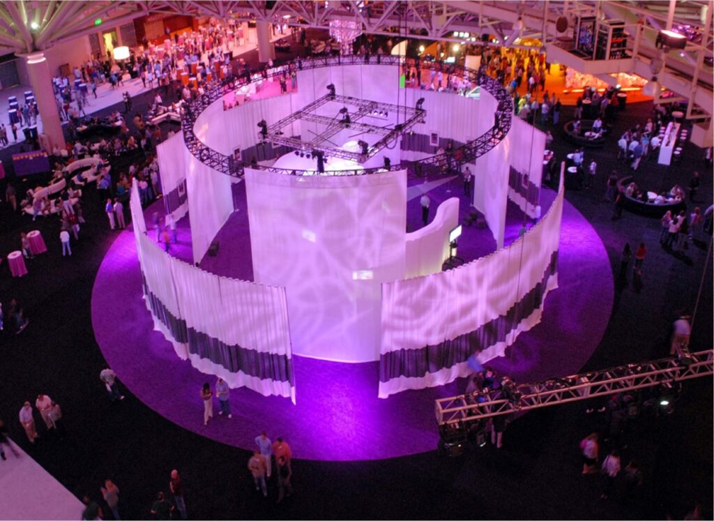 An overhead view of a vibrant event with modern lighting. Circular white structures with purple lighting dominate the scene. Many people are visible, mingling and exploring the various booths and displays in the spacious venue.