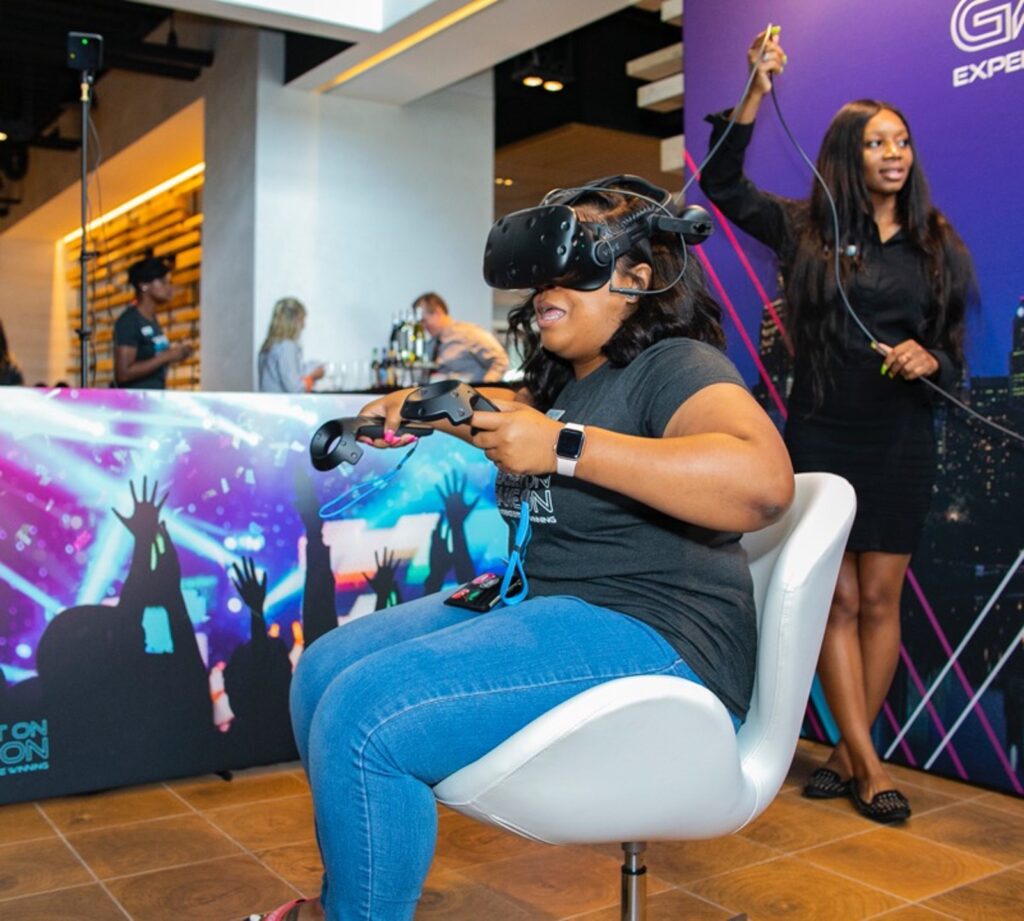 A person wearing a VR headset sits on a chair, holding controllers, immersed in a virtual experience. In the background, another person is standing and smiling, with a colorful banner featuring silhouetted concert-goers.