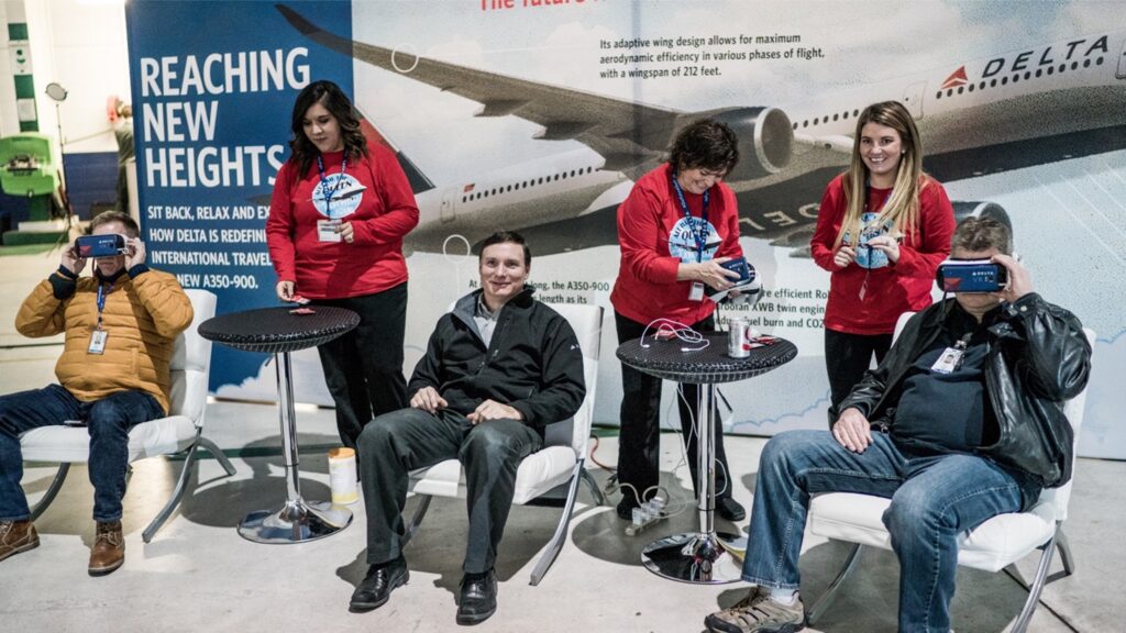 Five people seated, four using VR headsets. Three women wear matching red shirts and assist with devices. One man in a black jacket smiles, not wearing a headset. Behind them is a large aviation-themed banner.