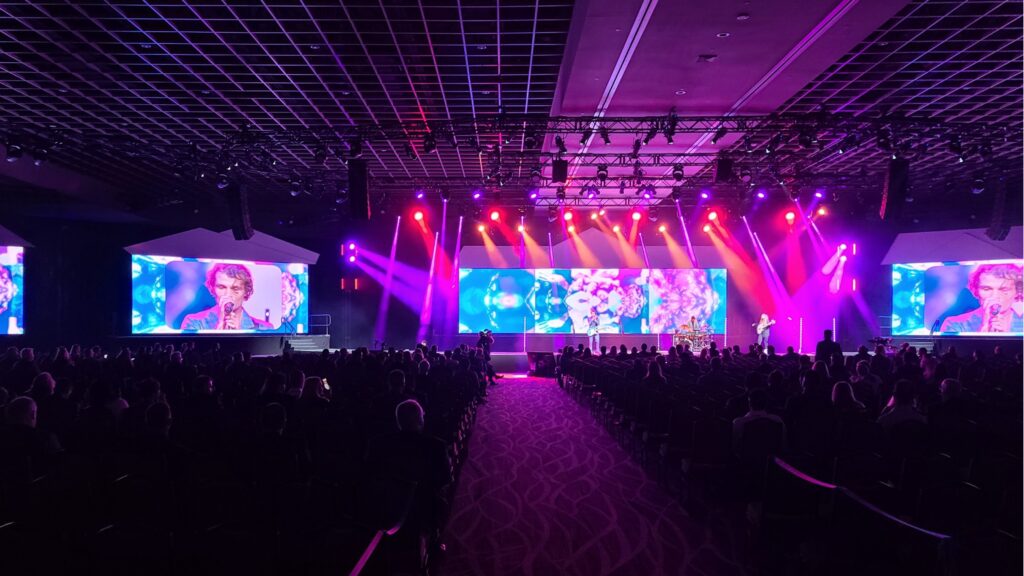 A large venue with vibrant purple and pink stage lighting features a musician on stage. Multiple screens display the performer to an audience seated in rows, creating a lively concert atmosphere.