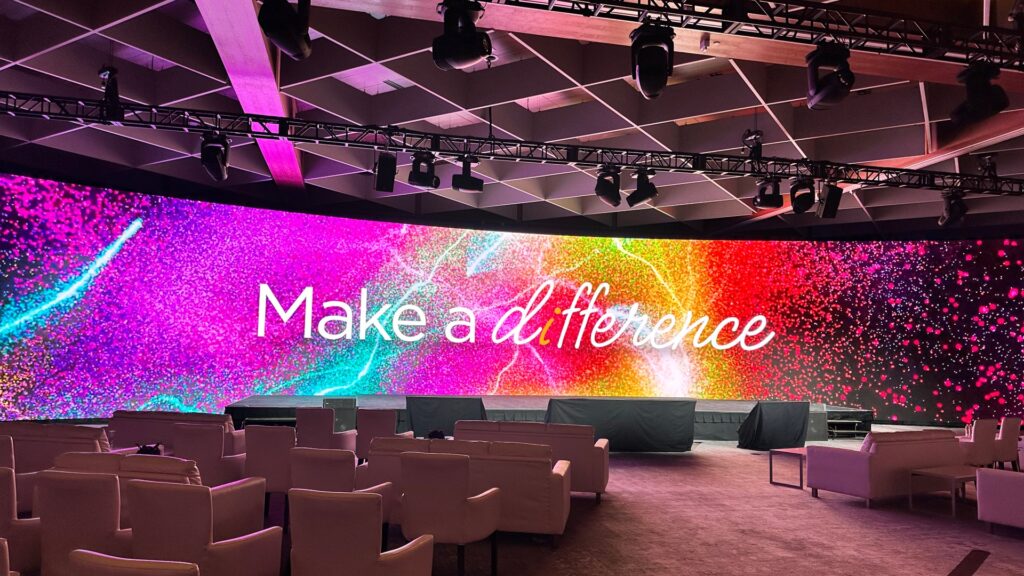 Large LED screen displays Make a Difference in elegant script over a vibrant, multicolor background. White chairs are arranged in rows facing the screen, under a ceiling with spotlights and beams.