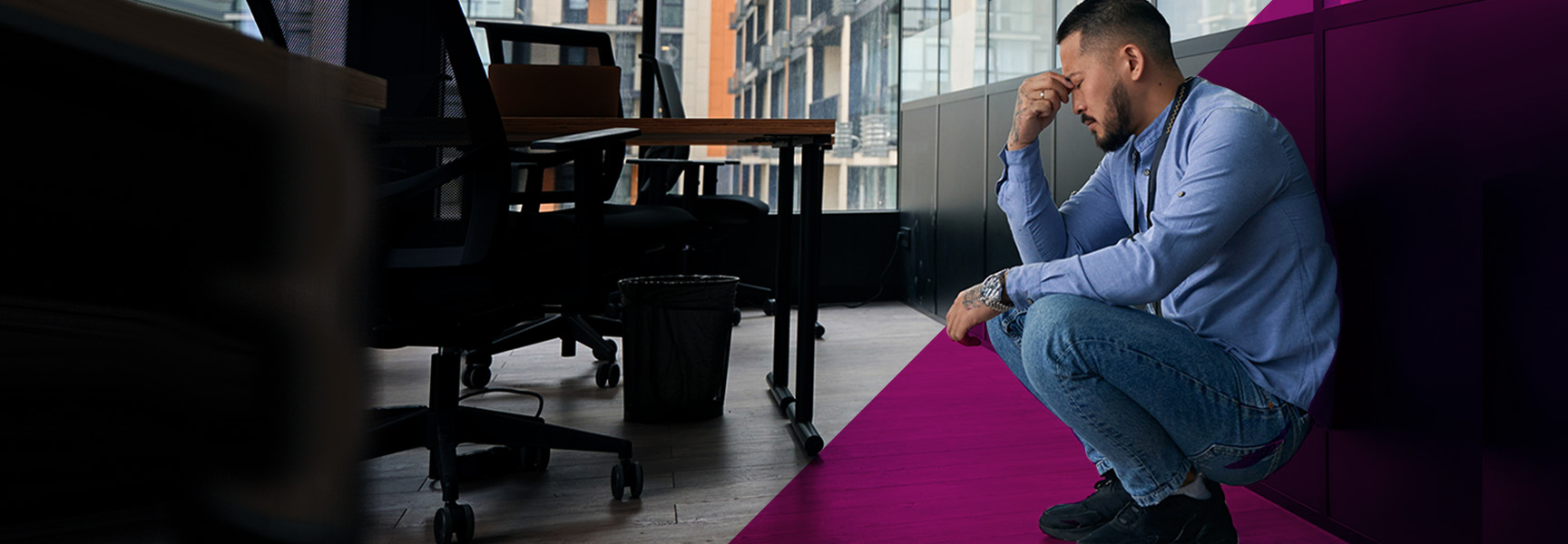 A person in a light blue shirt and jeans crouches in an office setting, hand on forehead, reflecting thoughtfully on potential mental health issues. The room features office chairs, desks, and large windows that add to the contemplative atmosphere.