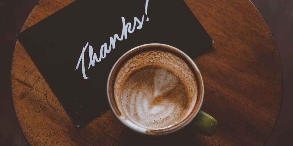 A cup of coffee with latte art sits on a wooden surface next to a black card with the word Thanks! written in white script.