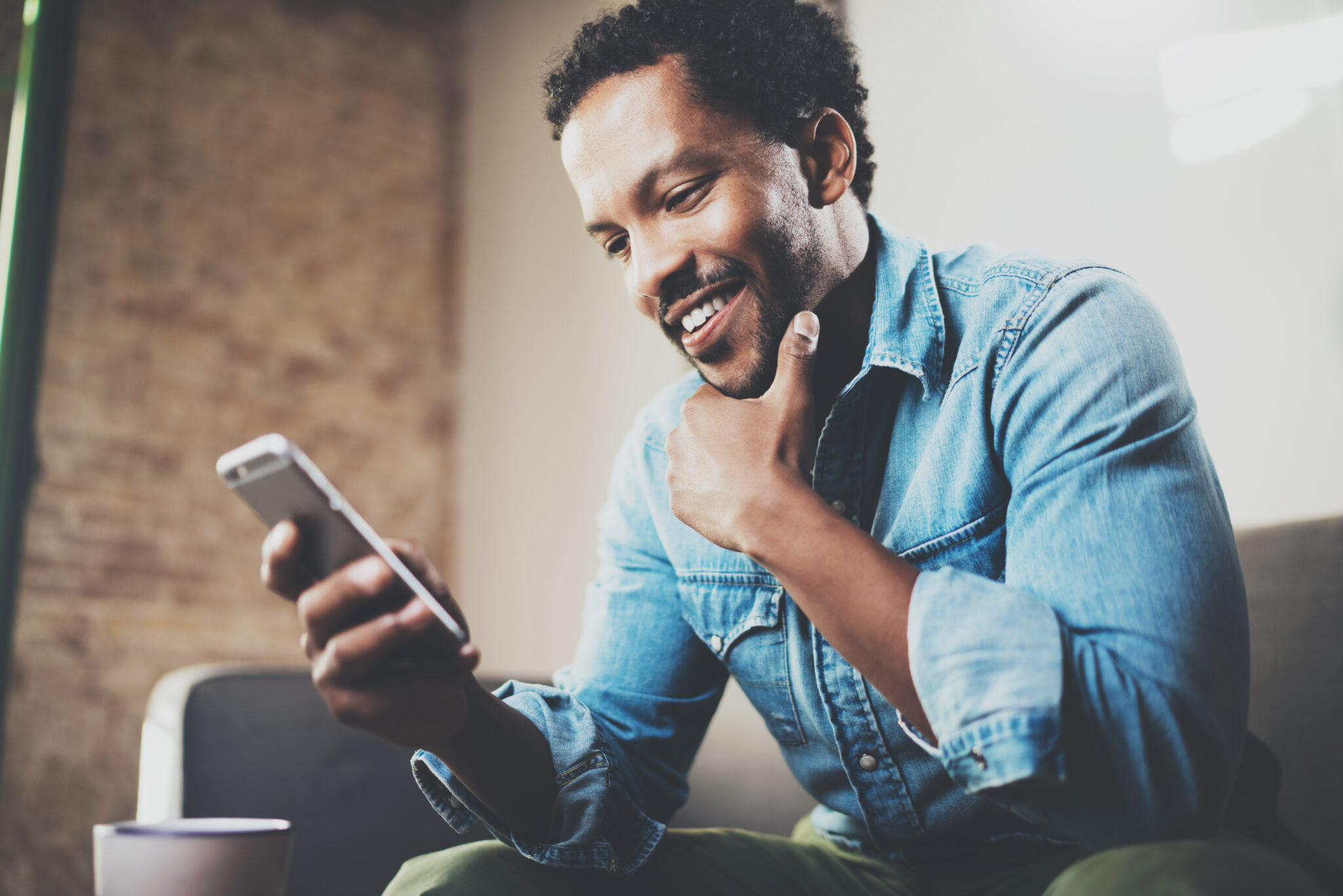 A smiling man in a denim shirt sits on a couch, looking at his smartphone with his hand on his chin, appearing happy and engaged—capturing a moment of employee engagement. A blurred cup is visible in the foreground.
