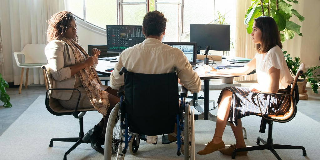 Three people, including a man in a wheelchair, are having a discussion in a bright office with large windows, desks, and computer monitors. The atmosphere appears collaborative and professional.