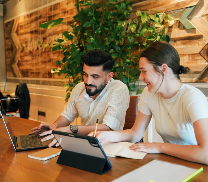 Two young adults sit at a wooden table, working together with a laptop, tablet, and notebook. They appear engaged and focused, with a green plant and wood-paneled wall in the background.