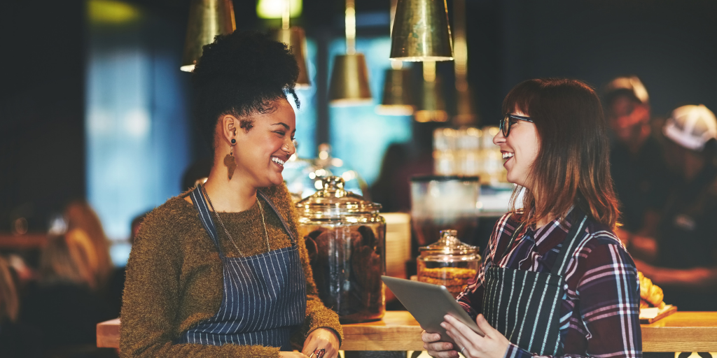 Two women wearing aprons stand behind a counter in a cozy cafe, smiling and talking as they discuss channel partner relationships. One holds a tablet, with jars of baked goods and warm lighting visible in the background.