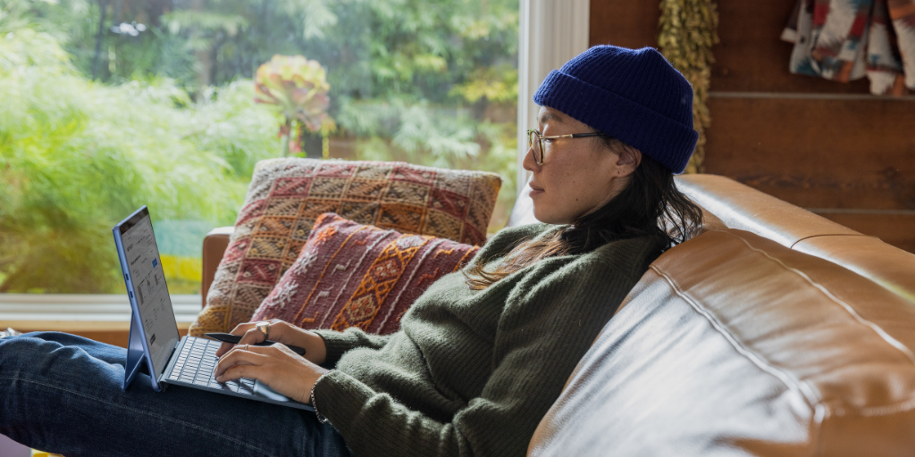 A person wearing glasses and a blue beanie sits on a couch using a laptop, with colorful pillows behind them and a large window showing greenery in the background.